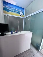 Um balcão de atendimento com divisória de vidro, um monitor de computador e uma pequena caixa azul com texto.
