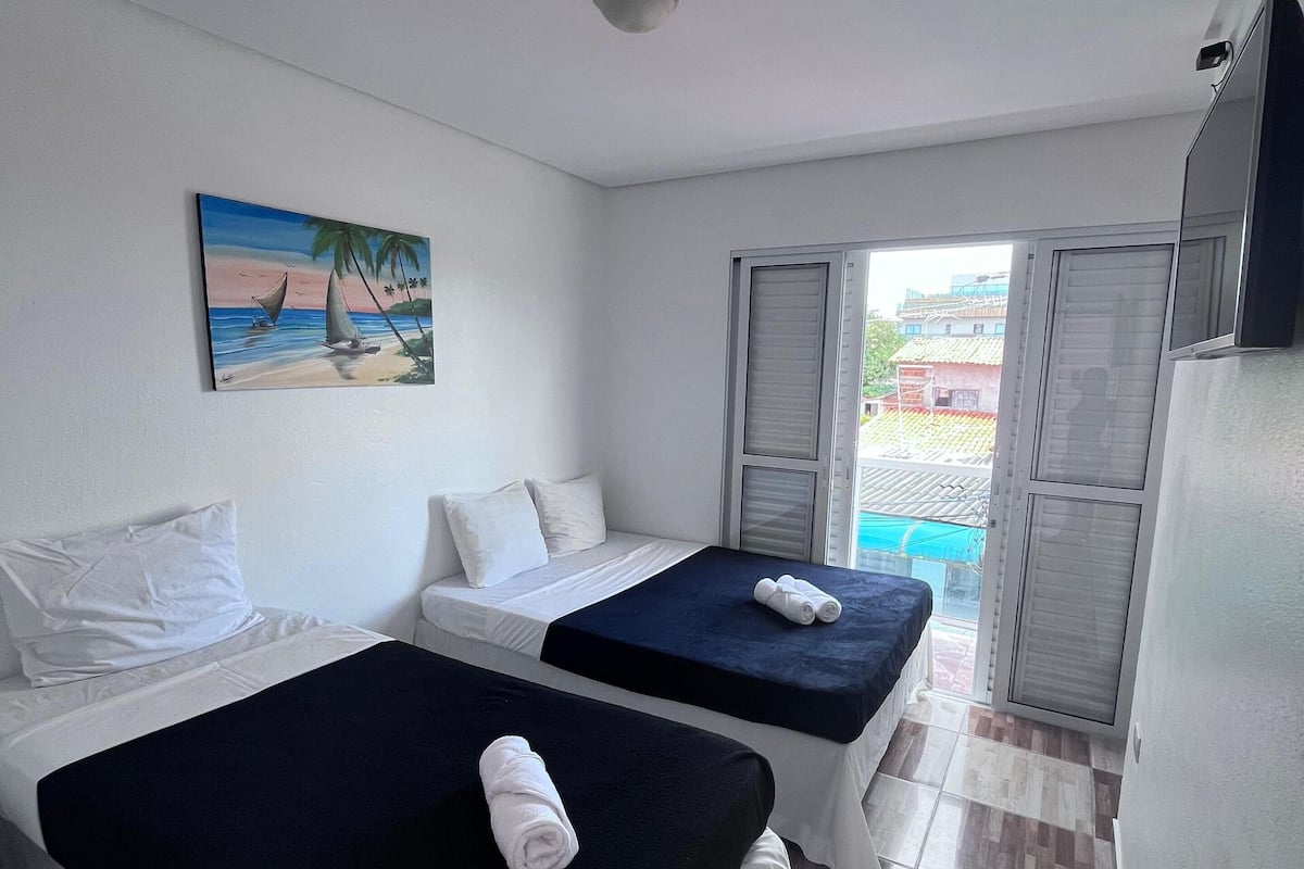 A hotel room with two beds, a painting of a beach scene, a window with a view of buildings, and a door leading to a balcony.