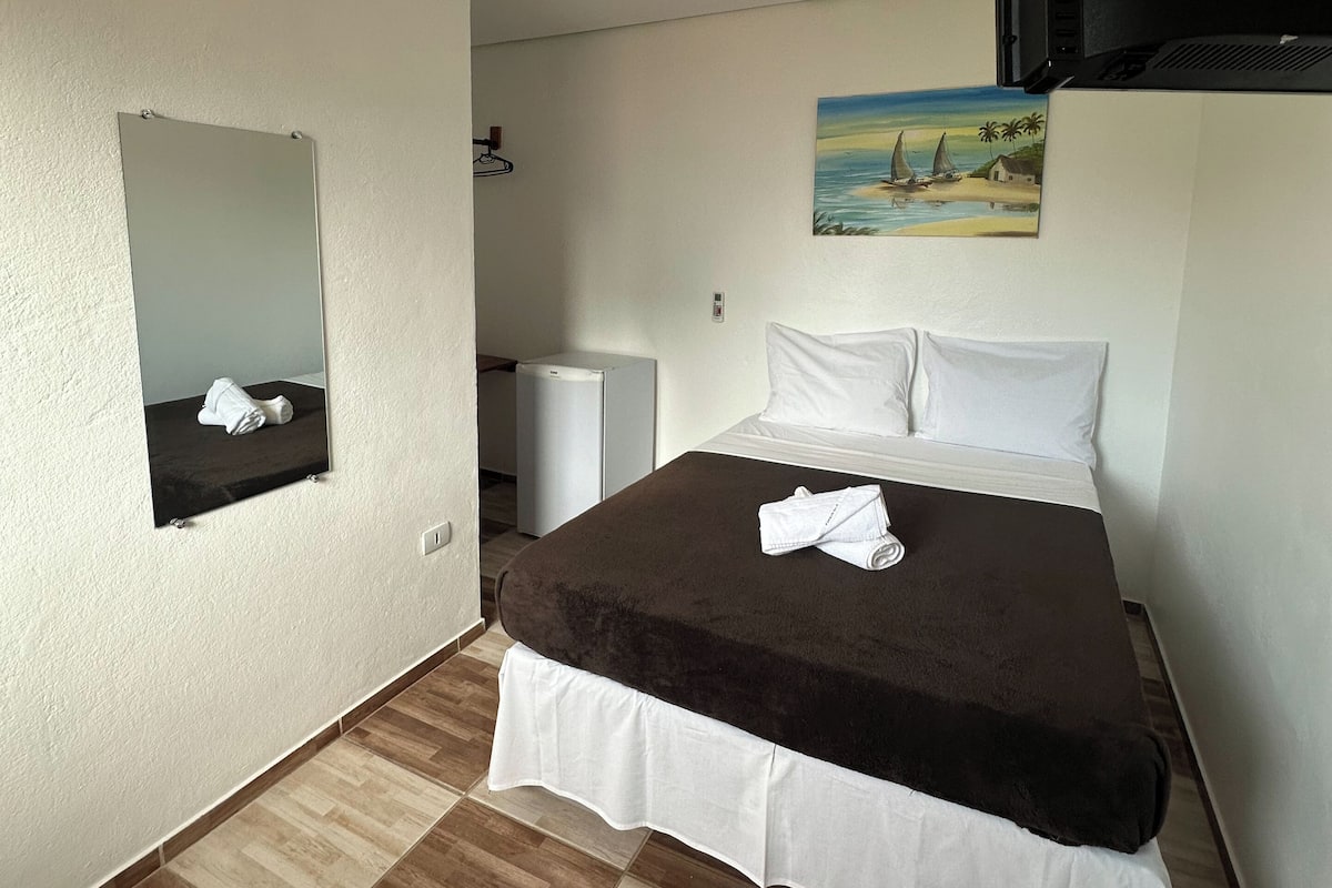 A hotel room with a bed, a mirror, a painting, a heater, and a television.