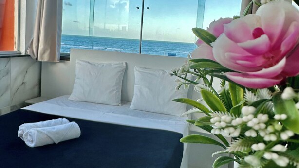 A hotel room with a bed, white pillows, a black bedspread, a white vase with flowers, and a view of the ocean through large windows. - Pousada Porto Navegantes (Ipojuca)