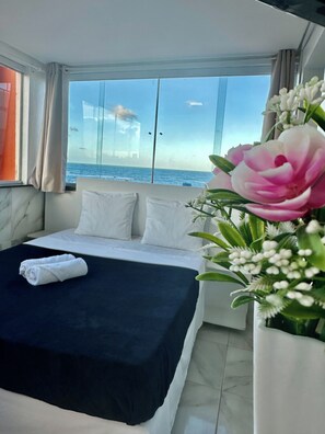 Une chambre d’hôtel avec un lit, des oreillers blancs, une couvre-lit noire, un vase blanc rempli de fleurs et une vue sur l’océan par de grandes fenêtres.