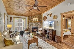A cozy living room with a stone fireplace, a ceiling fan, and a television.