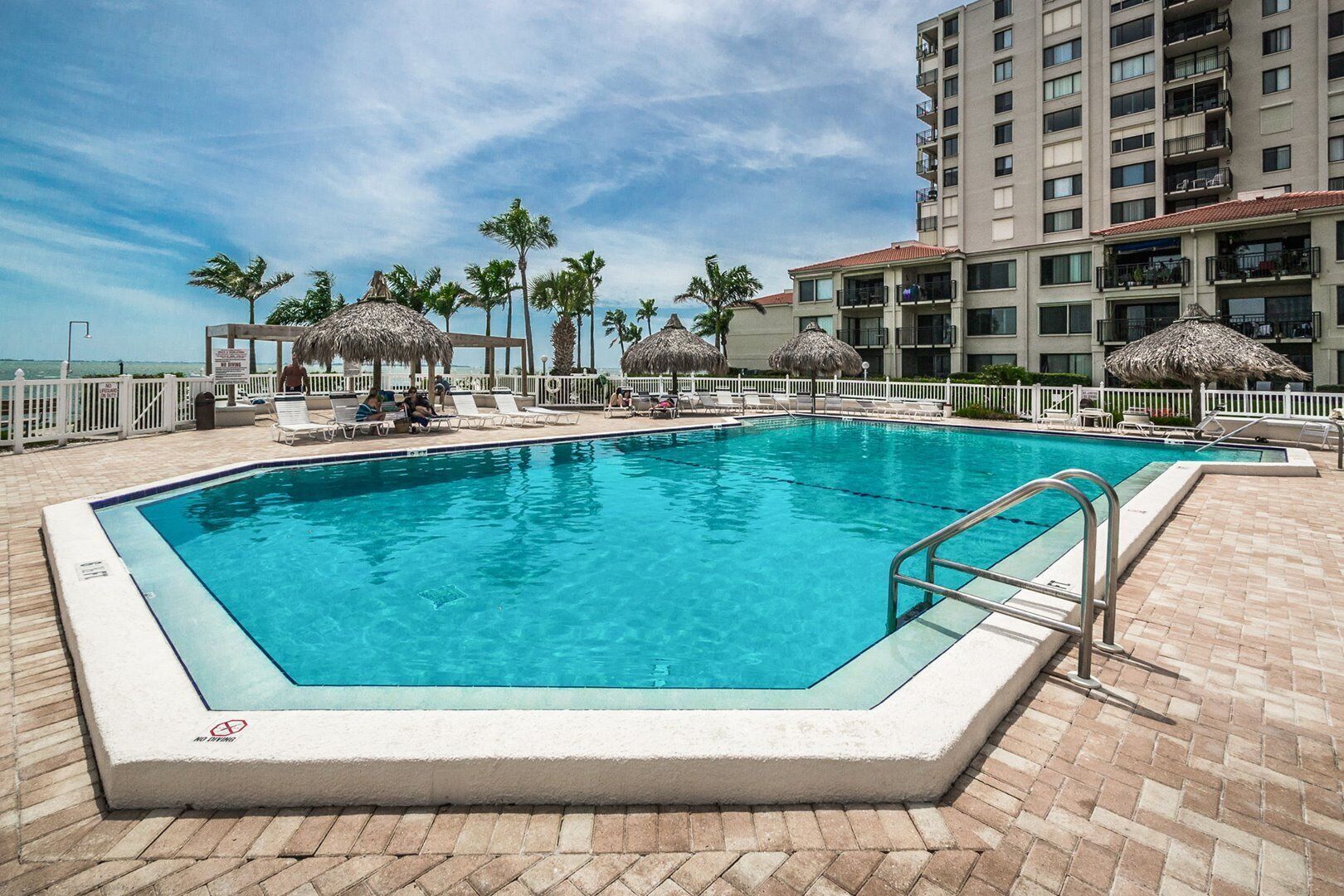 Una piscina rectangular con una escalera metálica, rodeada de sillas de descanso y palmeras, con un rascacielos al fondo.