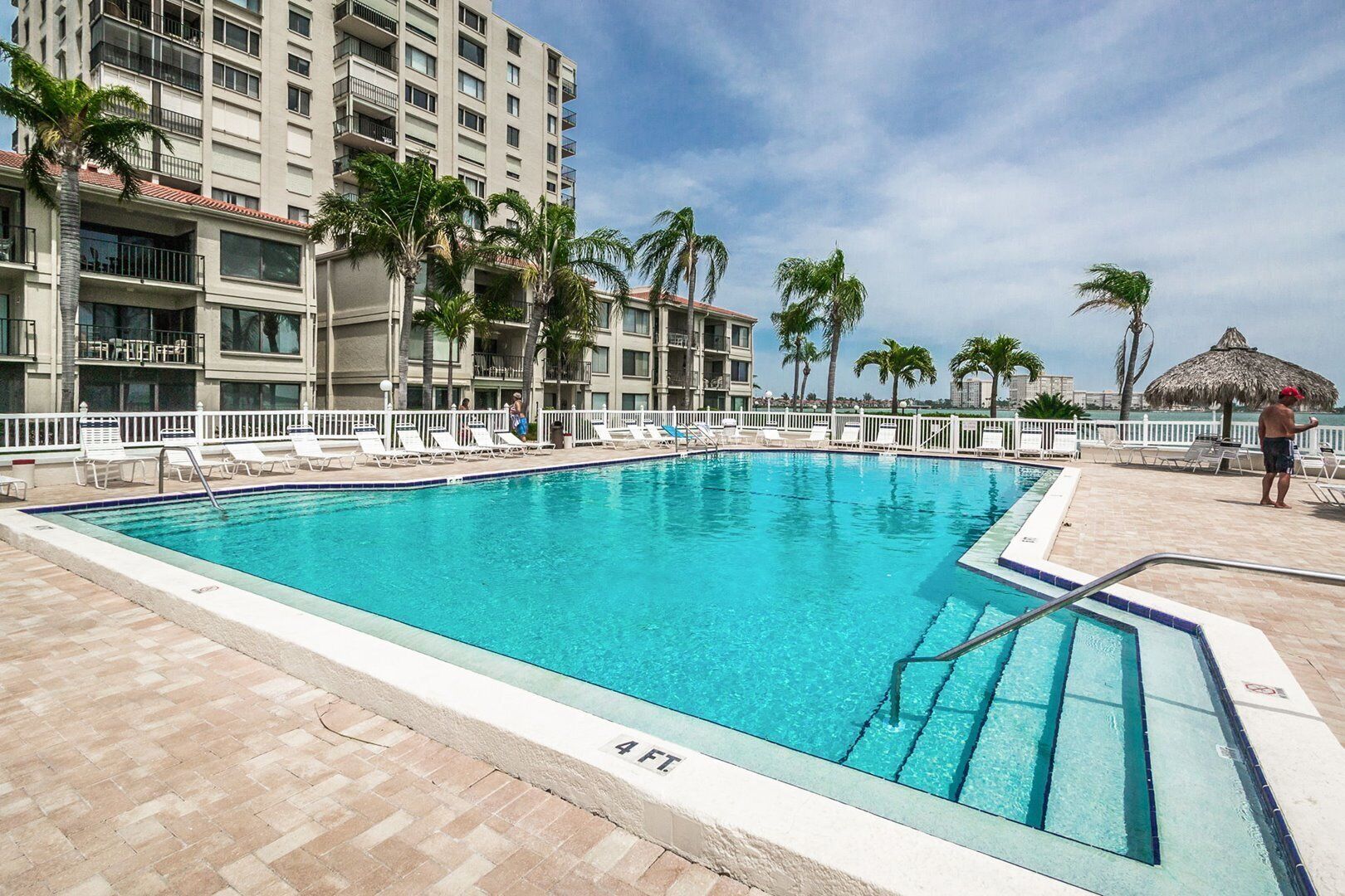 Une piscine dont la profondeur est indiquée à 1,20 mètre, entourée de chaises longues et de palmiers, avec un bâtiment en arrière-plan.
