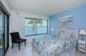 A bedroom with a large window, a bed with floral bedding, a nightstand with a lamp, and a chair by the window. - Discover the Epitome of Waterfront Living in this Stunning condo on Isla del Sol!~ (St Petersburg)