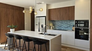 A modern kitchen with a central island, bar stools, a stainless steel refrigerator, a built-in oven, and a blue brick backsplash.