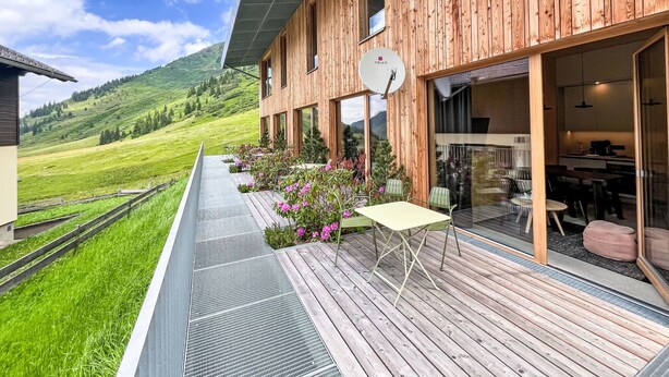 A modern wooden building with large windows, a balcony with a table and chairs, and a view of a green valley.
