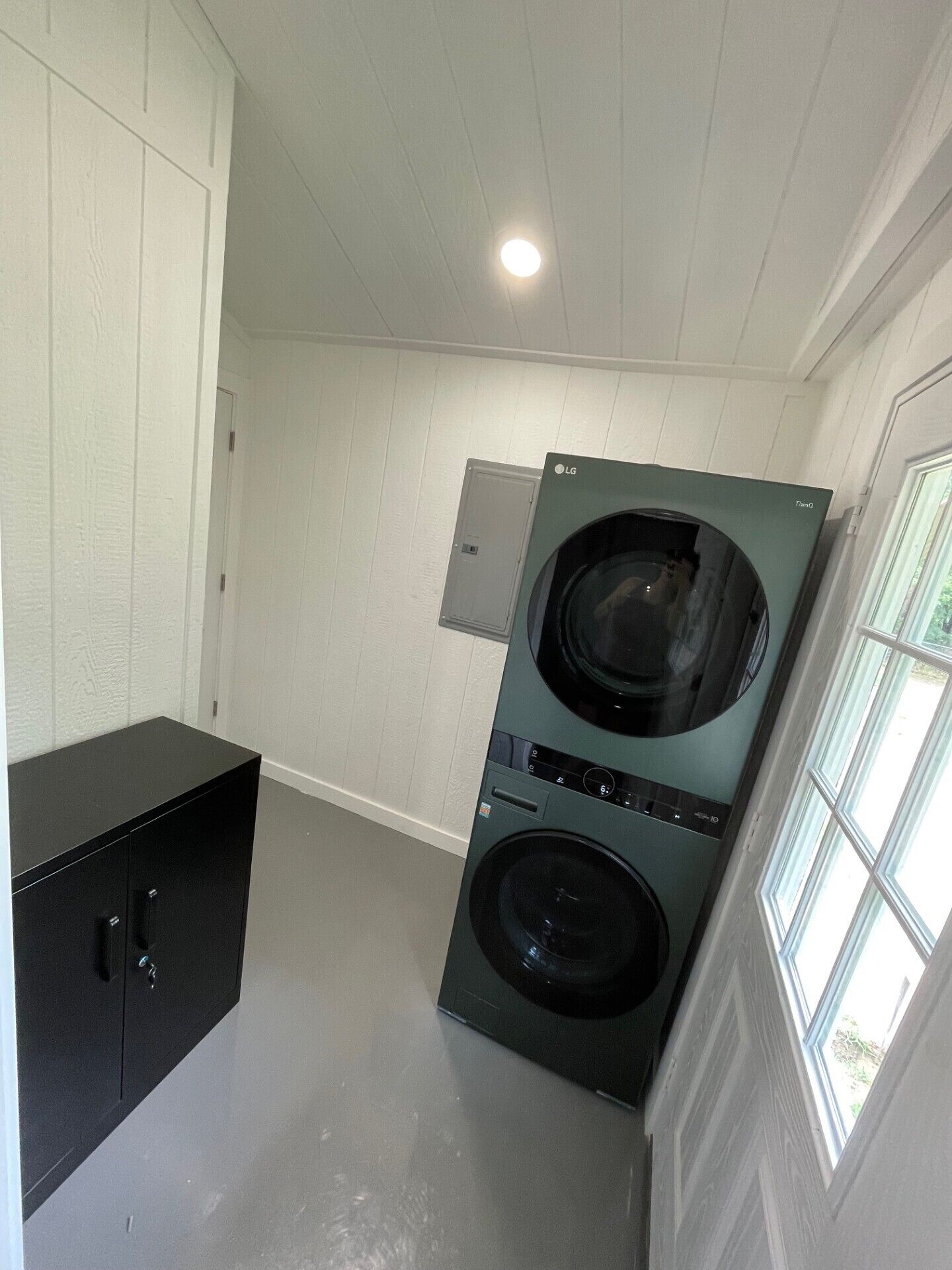 A laundry room with a front-loading washer and dryer, a black storage cabinet, and a window providing natural light.