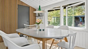 Un'area pranzo moderna con un tavolo rotondo, sedie bianche, una lampada a sospensione verde e vista su piante.