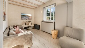Ett mysigt vardagsrum med en soffa, ett träbord, en TV-bänk med böcker och en platt-TV, ett fönster med utsikt över grönområden och en rustik träpall.