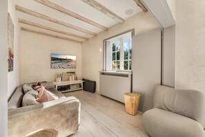 Un salon chaleureux avec un canapé, une table basse en bois, un meuble pour la télévision agrémenté de livres et d’un téléviseur à écran plat, une fenêtre donnant sur un paysage verdoyant et un tabouret en bois rustique.