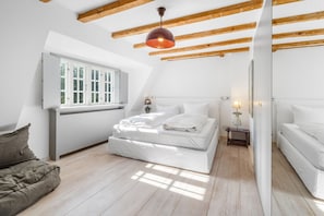 Ein Schlafzimmer mit schrägen Wänden, einem Bett mit weißer Bettwäsche, einem Nachttisch mit Lampe und einem Fenster mit weißen Fensterläden.