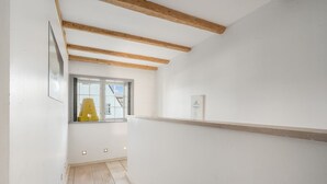 Een smalle gang met houten balken in het plafond, een raam met een gele stoel erbuiten, en een witte muur met een lijstwerk afbeelding.