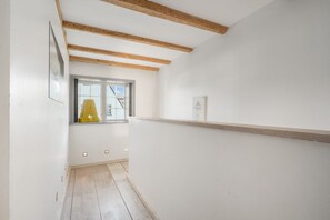Kapea käytävä, jonka katossa on puupalkkeja, ikkuna, jonka ulkopuolella on keltainen tuoli, ja valkoinen seinä, jossa on kehystetty kuva.