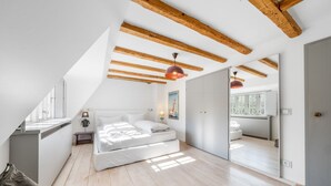 Een slaapkamer met een loft-plafond, houten balken, een bed met witte beddengoed, een nachtkastje, een lamp en een poster aan de muur.