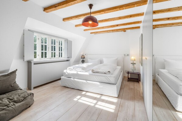 A bedroom with a sloped ceiling, a bed with white bedding, a bedside table with a lamp, and a window with white shutters.