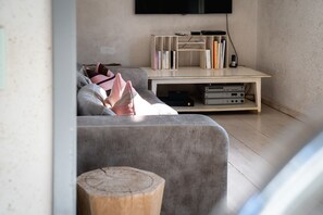 Ein Wohnzimmer mit grauem Sofa, einem Holztisch und einem Bücherregal mit Büchern und elektronischen Geräten.