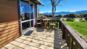 A wooden deck with a glass door leading to a room, a small table, and a chair. A tree is visible in the background with a mountain view. - Manapouri Holiday Park (Manapouri)