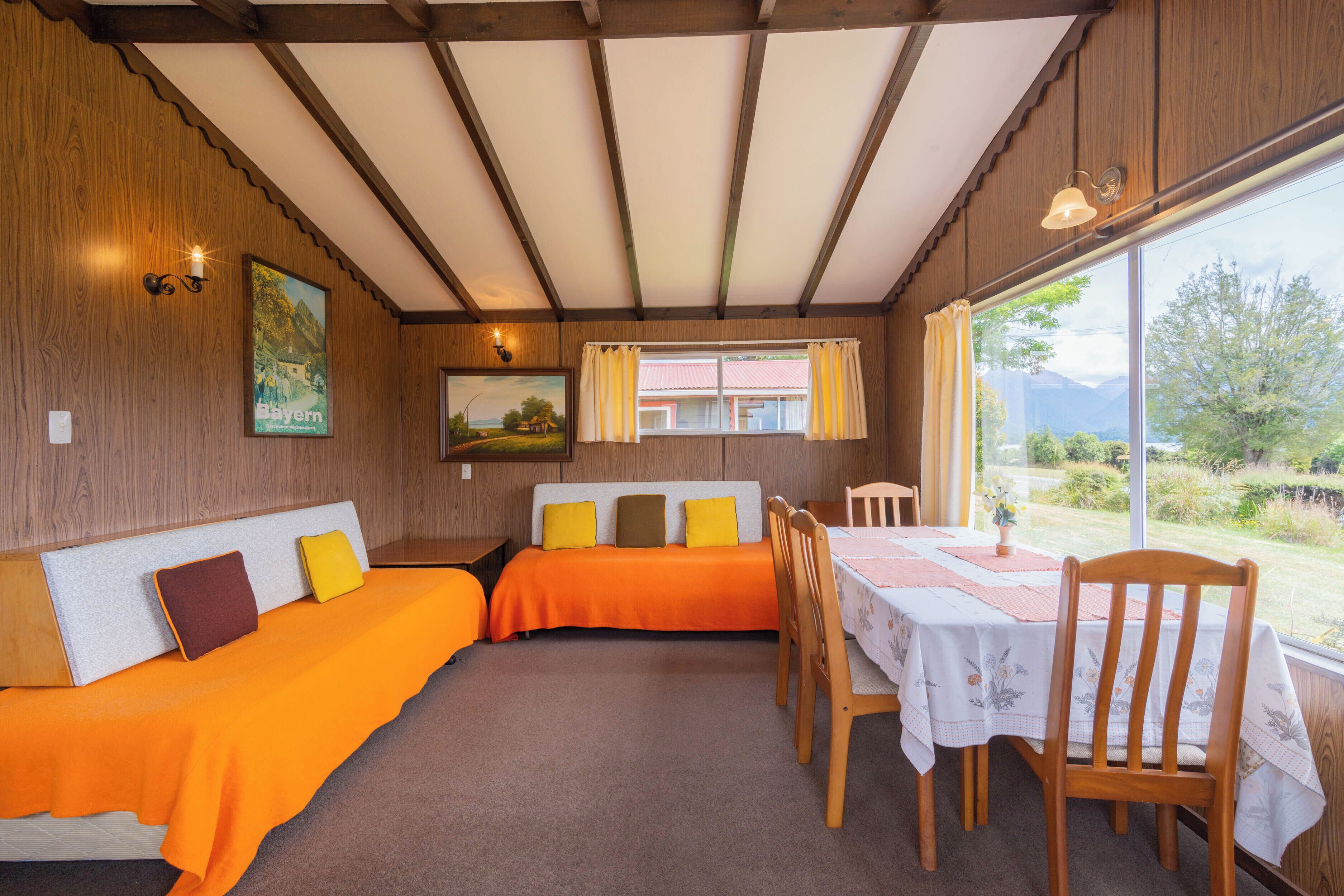 Una habitación con techo de madera, una cama con ropa de cama naranja, una mesa con mantel floral y vistas al exterior con vegetación.