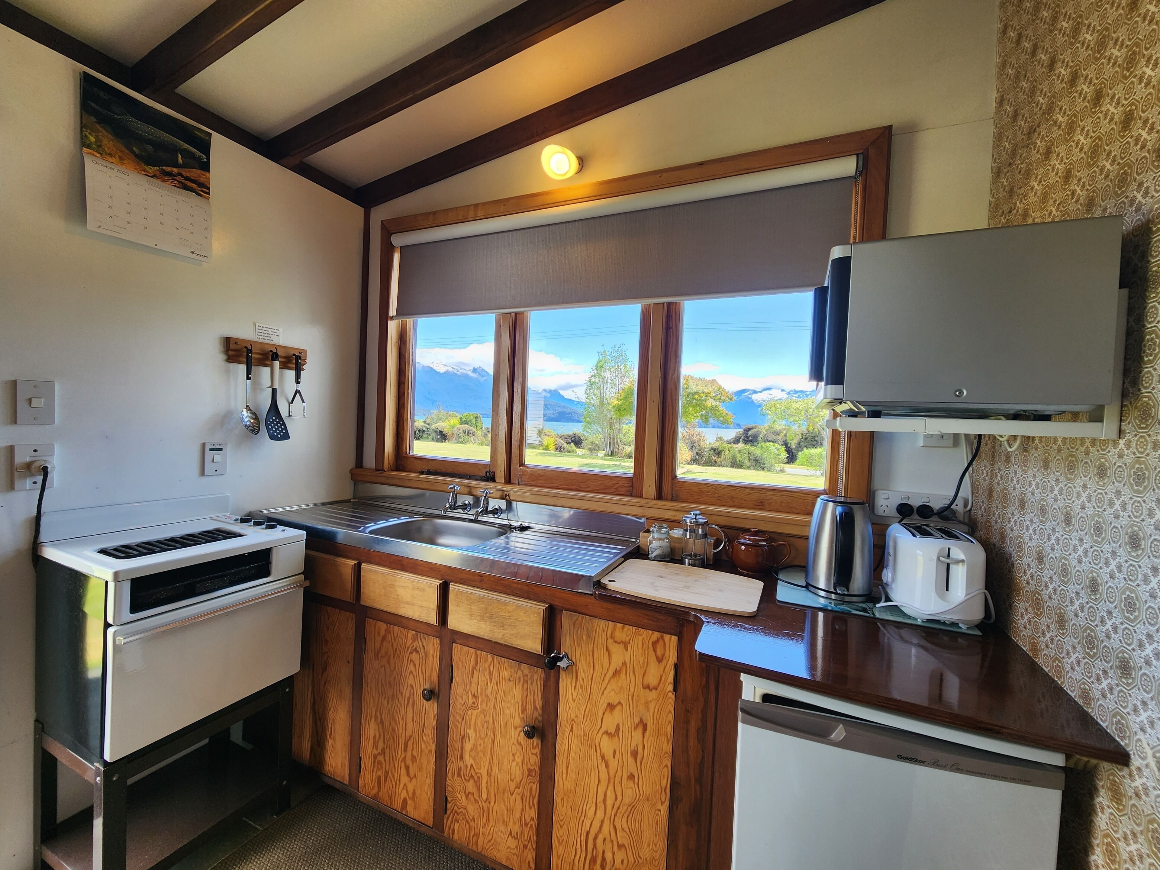 Una cocina compacta con armarios de madera, horno empotrado, lavamanos y ventana con vistas panorámicas a montañas y vegetación.