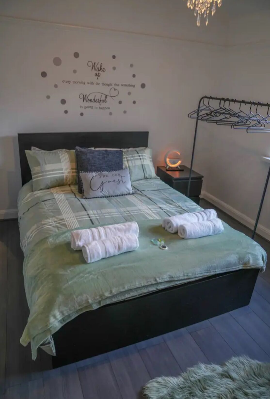 A neatly made bed with a green bedspread, multiple pillows, and rolled towels. There is a wall decoration with motivational phrases and a wall-mounted light fixture.
