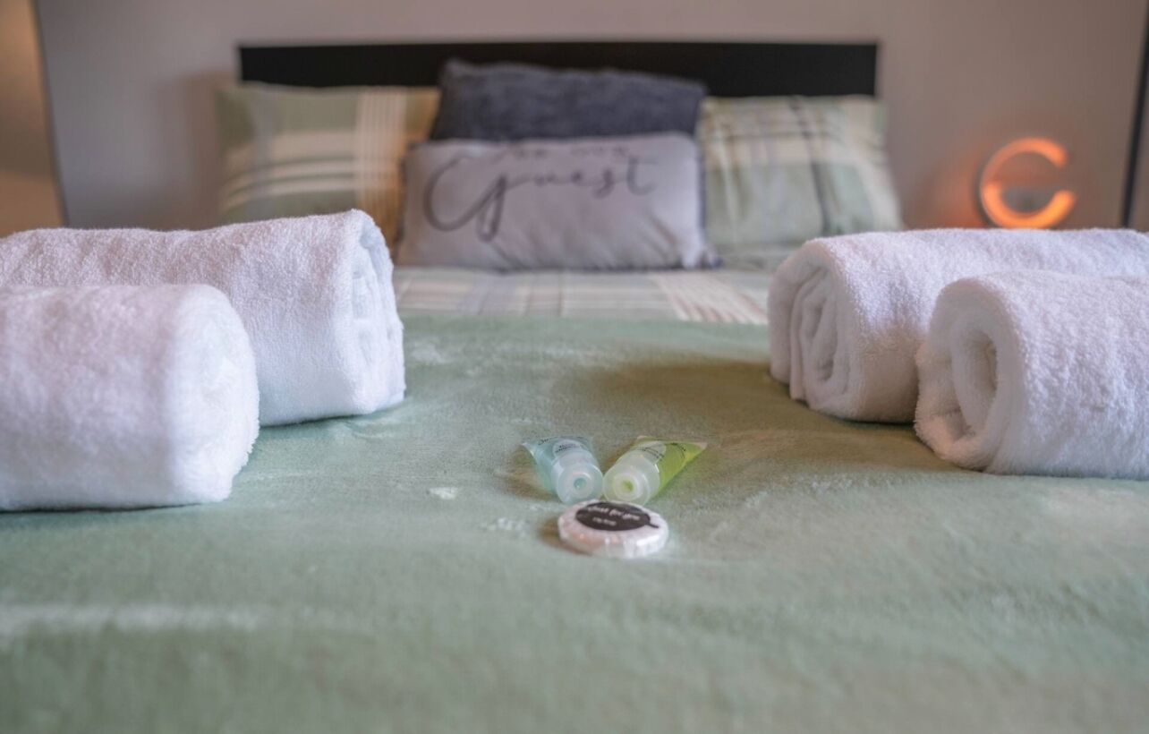 A neatly made bed with rolled white towels, a green blanket, and a small bottle of lotion on the bed.