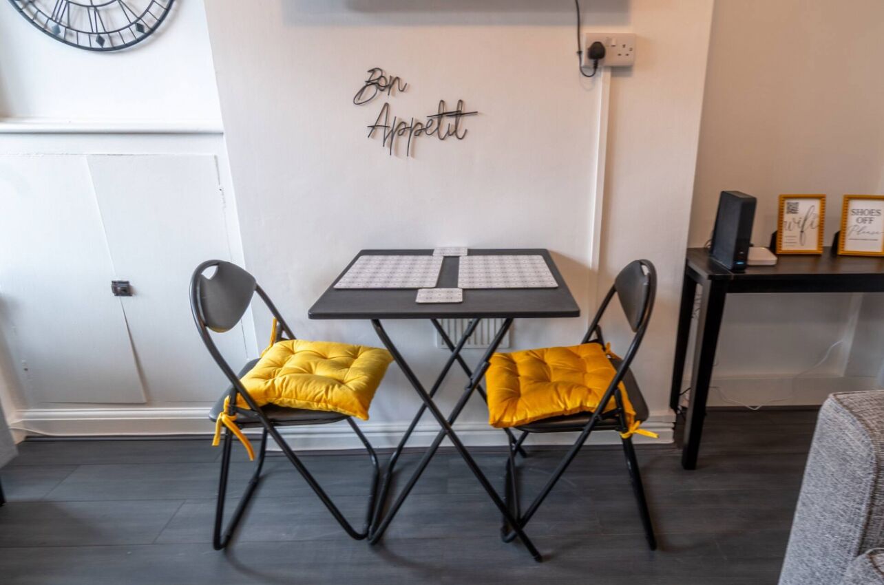 Ein kleiner Raum mit einem Tisch und zwei Stühlen, einer Uhr an der Wand und einem Schild mit der Aufschrift 'Bon Appetit'.