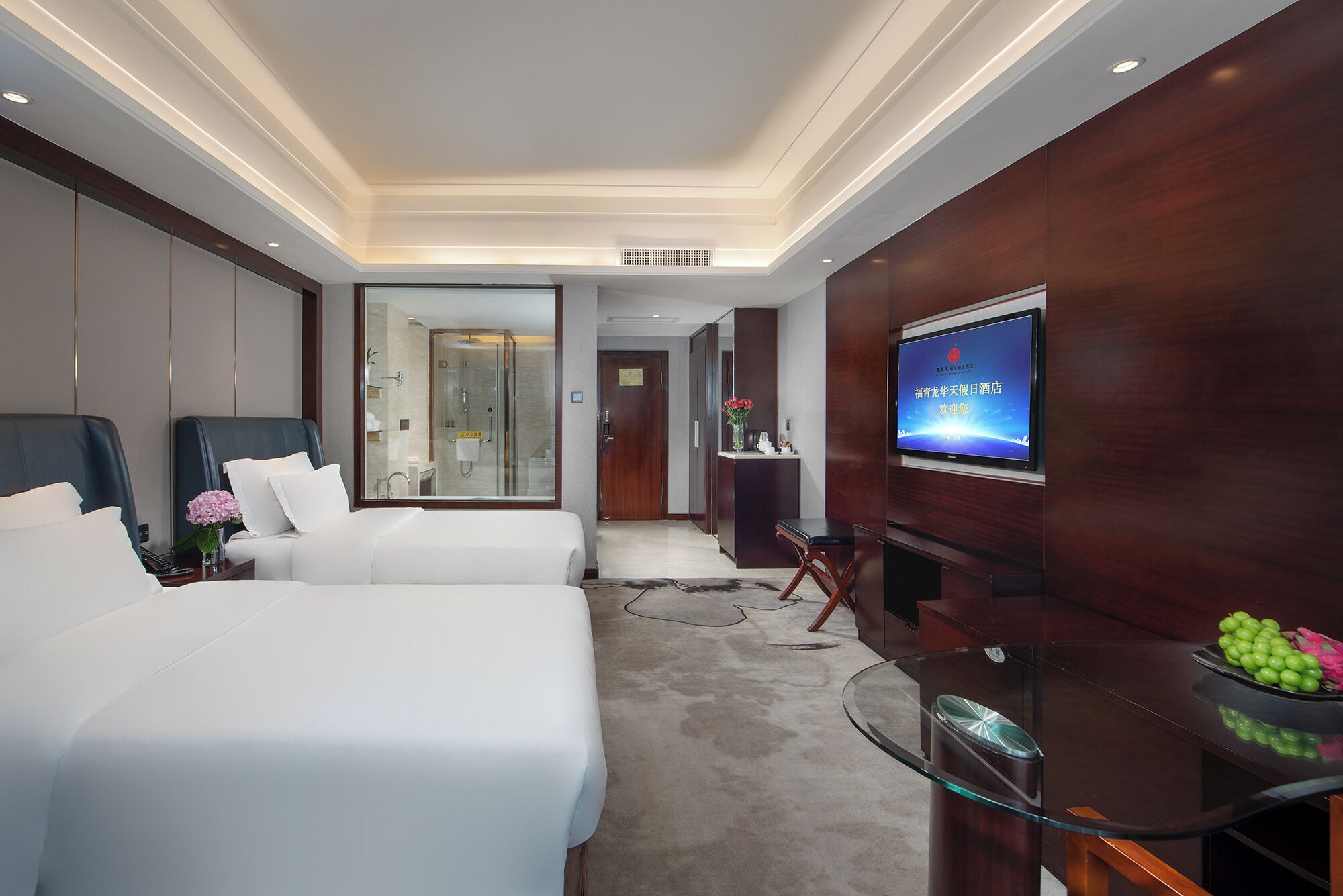 Habitación de hotel con dos camas, un televisor, un escritorio y un baño visible a través de una puerta de vidrio.