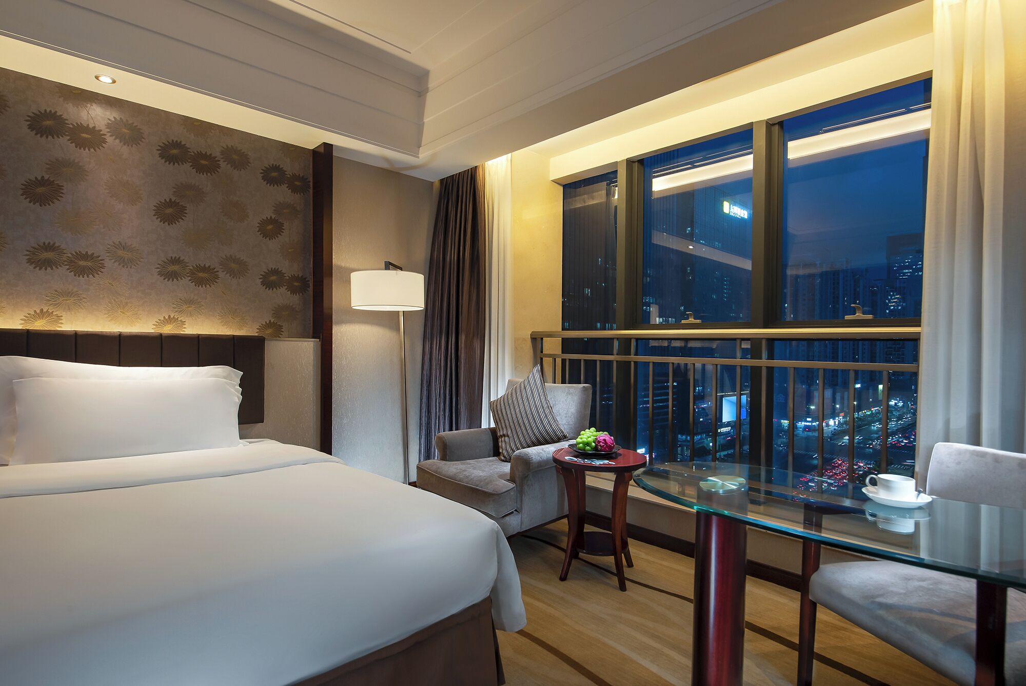 A hotel room with a bed, a desk with a chair, a lamp, and a view of the city at night.