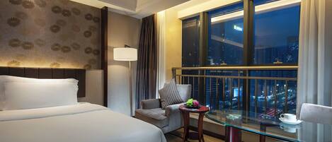 A hotel room with a bed, a desk with a chair, a lamp, and a view of the city at night.