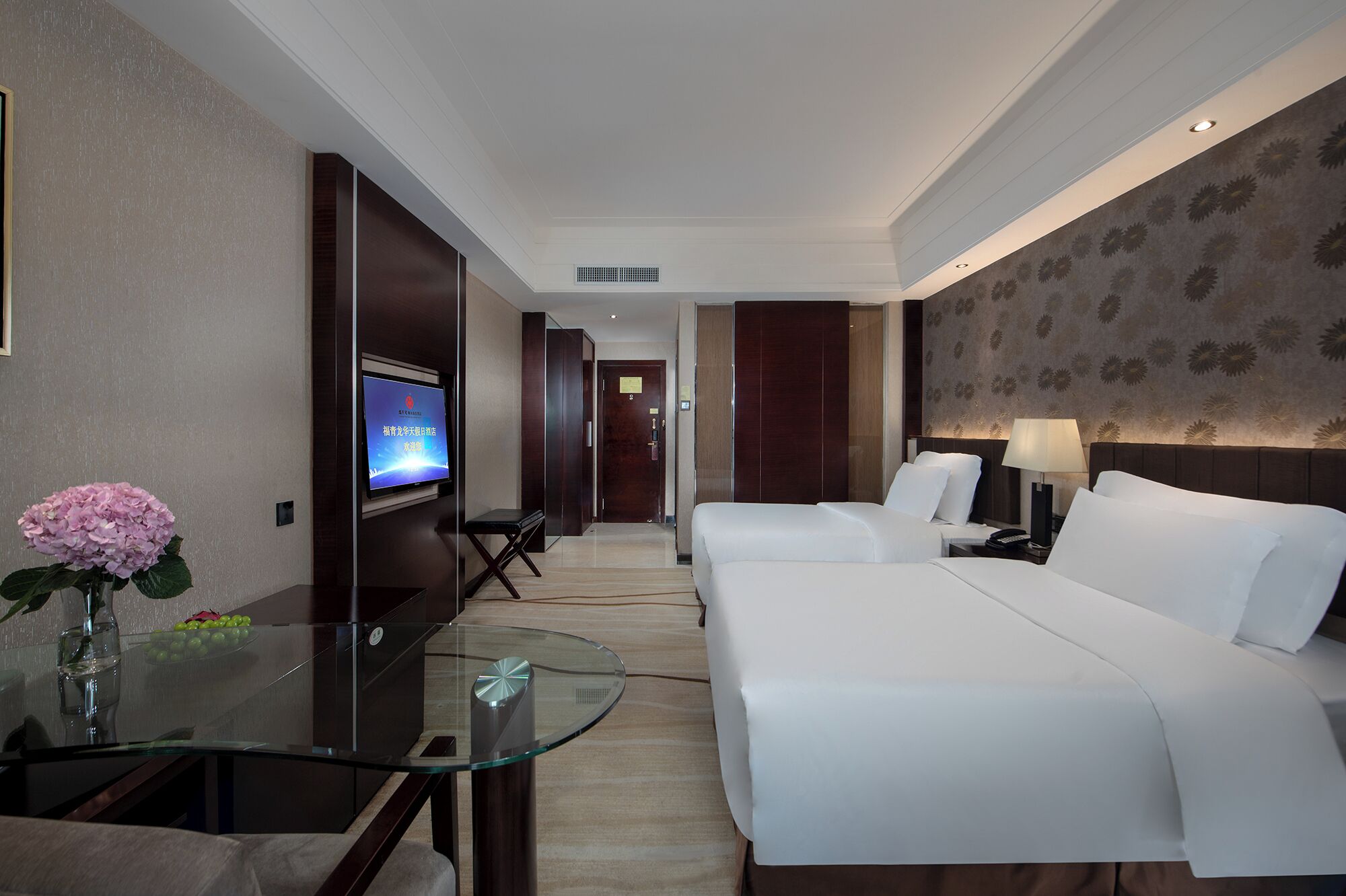 Habitación de hotel con cama, mesitas de noche, televisión y una mesa de vidrio con un florero.