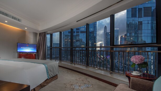 A hotel room with a large window offering a city view, a bed, a TV, and a small table with a vase. - Fuqinglong Huatian Holiday Hotel (Shenzhen)