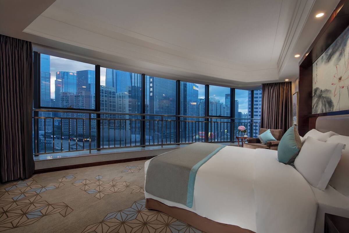 Een hotelkamer met een groot bed, een nachtkastje en uitzicht op de stadshemel door grote ramen.