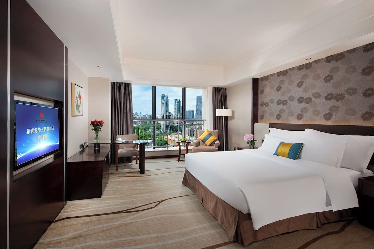 Een hotelkamer met een groot bed, een bureau met stoel, een televisie en uitzicht op de stad door grote ramen.