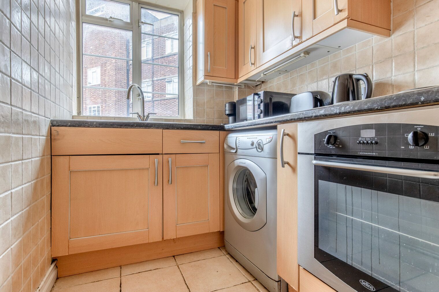 Een keuken met houten kastjes, een wasmachine, een fornuis en een raam met uitzicht op gebouwen.
