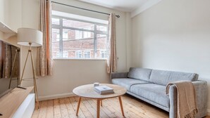 Een moderne woonkamer met een grijze bank, een ronde houten salontafel, een vloerlamp en een raam met gordijnen.