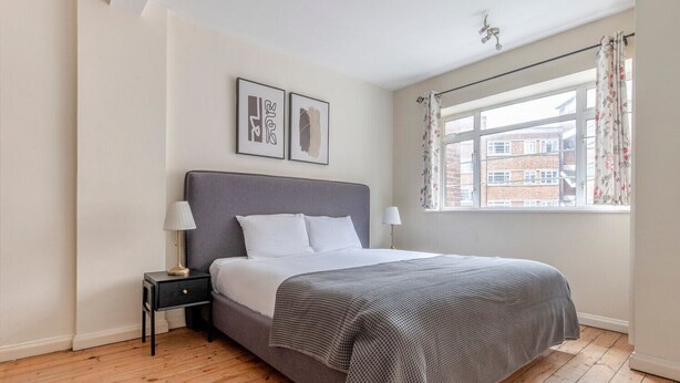 Een slaapkamer met een grijs bed, witte kussens, een grijze deken, een klein nachtkastje met een lamp, en een raam met bloemen gordijnen.