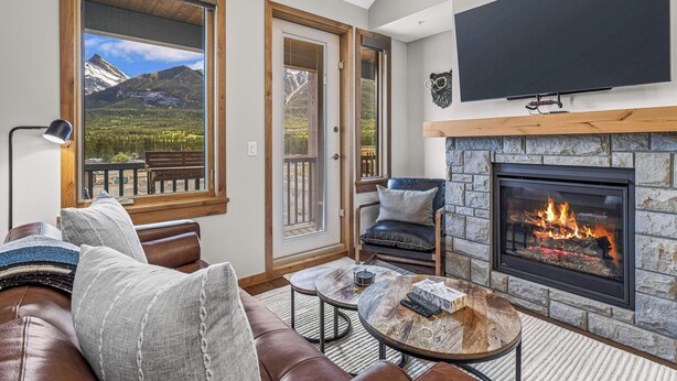 A cozy living room with a fireplace, comfortable seating, a large window with a mountain view, and a flat-screen TV mounted above the fireplace. - Spring Creek Vacations 1 BR Condo 311TM (Canmore)