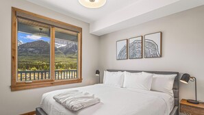 A bedroom with a large bed, a view of mountains through a window, and two framed artworks on the wall. - Spring Creek Vacations 1 BR Condo 311TM (Canmore)