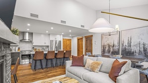 A modern kitchen with white cabinets, a stainless steel refrigerator, and a wooden dining area with bar stools. - Spring Creek Vacations 2 BR Condo 309TM (Canmore)