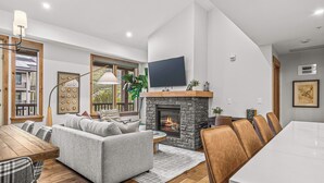A modern living room with a fireplace, a dining area, and a large TV mounted on the wall. - Spring Creek Vacations 2 BR Condo 309TM (Canmore)