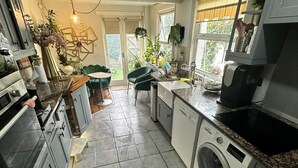 A compact kitchen with a washing machine, a coffee maker, and a dining area with a green chair. - My Sky Secret Garden Near atractions (London)
