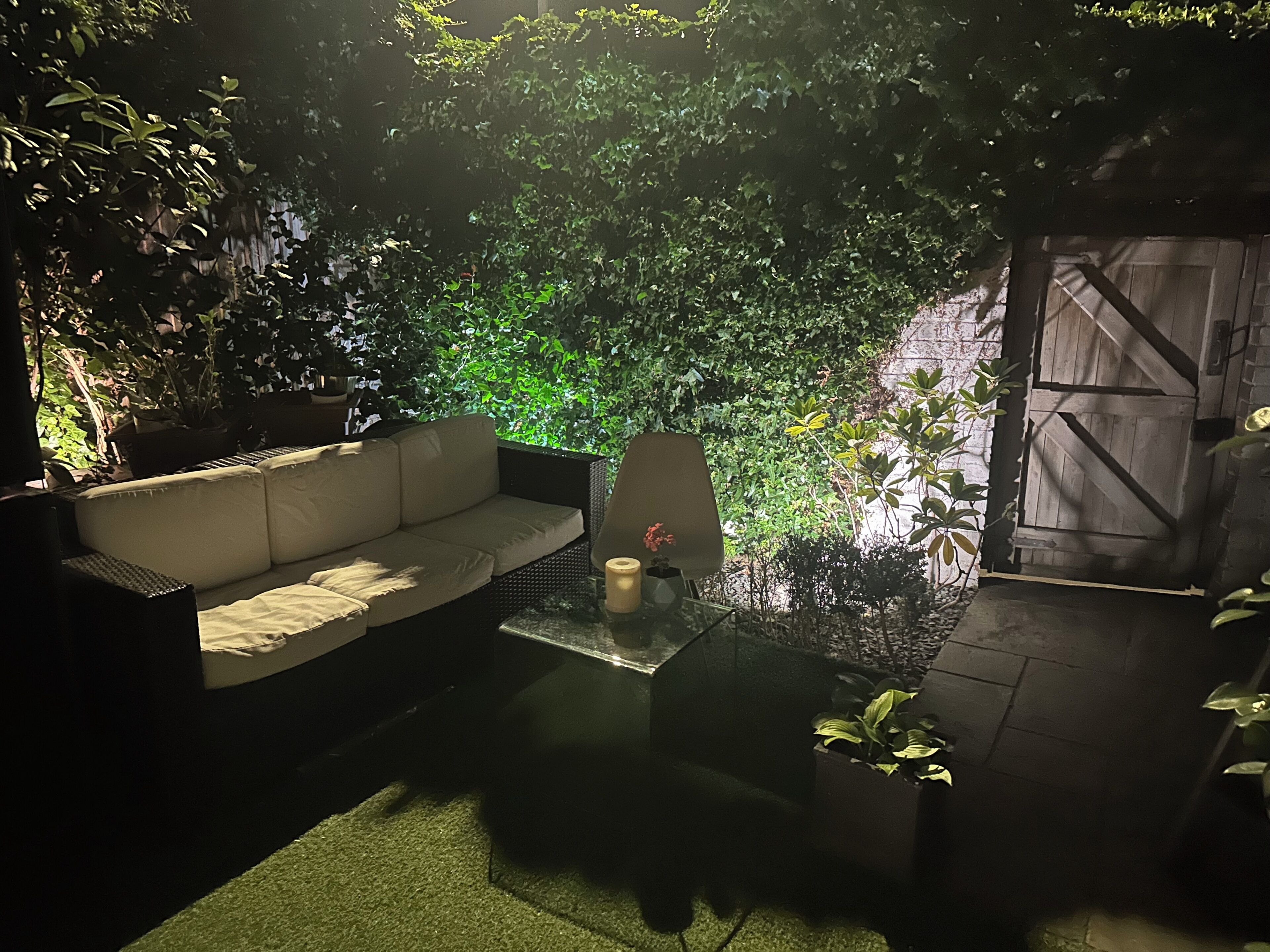 A cozy outdoor seating area with a sofa and a table, surrounded by lush greenery and a wooden gate.