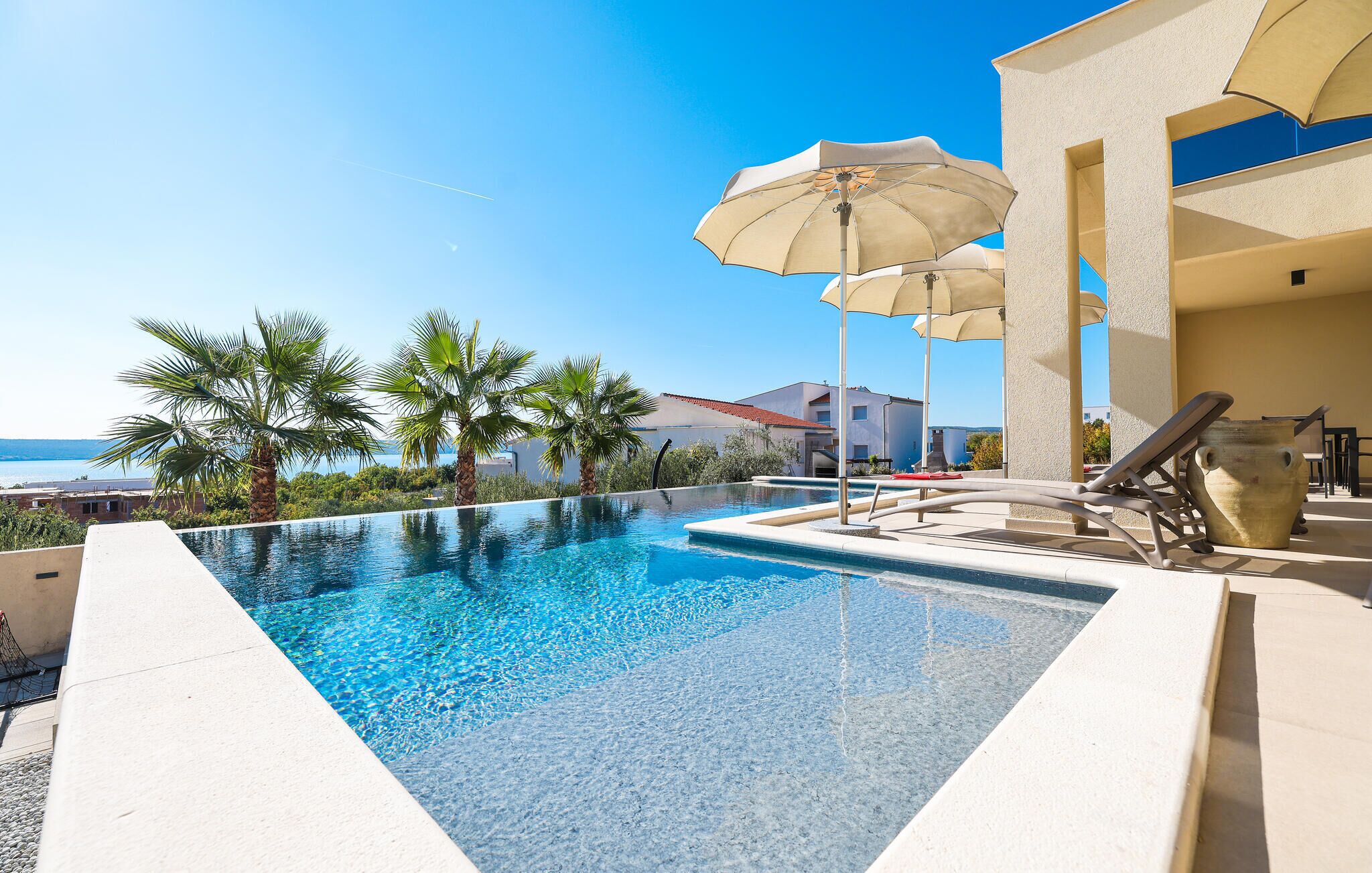 Ein Swimmingpool mit Liegestühlen und Sonnenschirmen, Palmen und Blick auf das Meer.