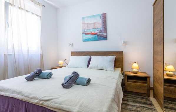 A neatly made bed with two pillows, two rolled towels, and a painting of a harbor scene on the wall.