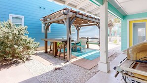 Een overdekt buitenterrein met een houten pergola, een tafel en stoelen, omringd door een blauw gebouw en een witte hekken.