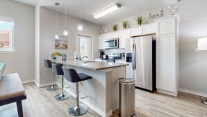 A modern kitchen with a central island, bar stools, a stainless steel trash can, and a refrigerator. - Shared Heated Pool, Golf Cart Access, Pet Friendly (Port Aransas)