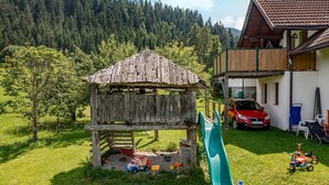 Una casita de juegos de madera con un tobogán, un coche rojo y una raqueta de tenis en un césped con montañas al fondo.