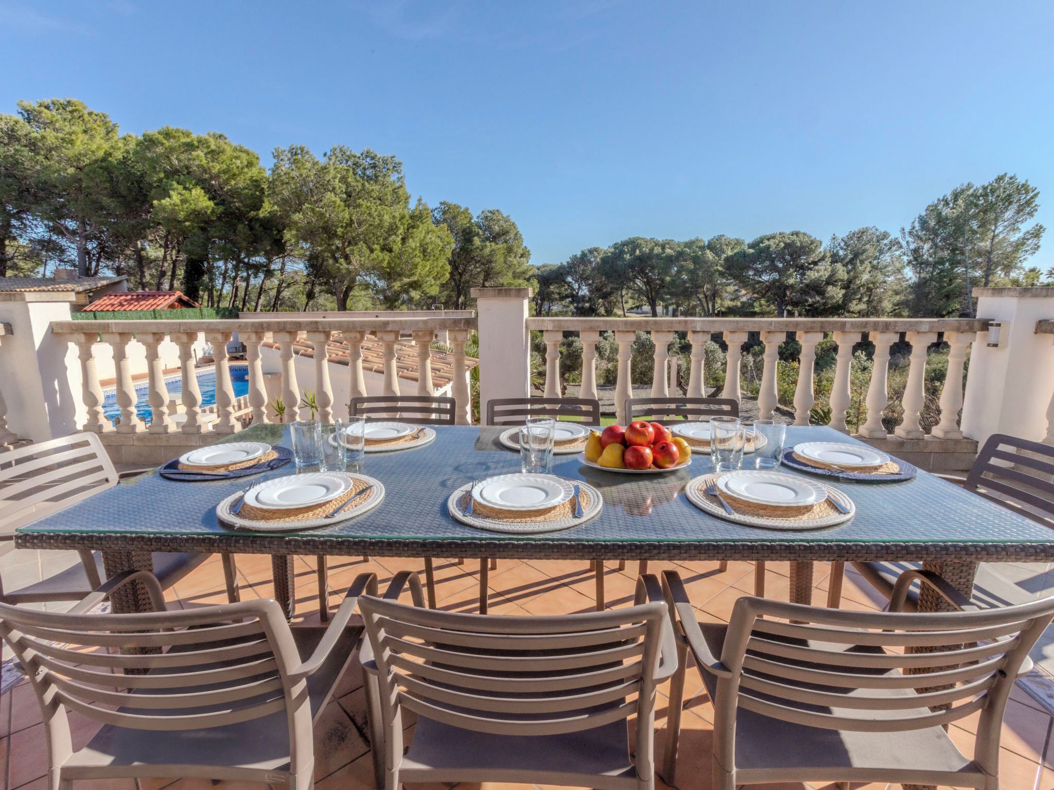 Un área de comedor al aire libre con una mesa lista para seis personas, rodeada de sillas, con vistas a una piscina y árboles.