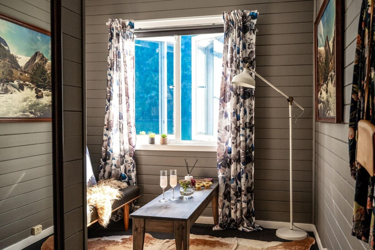 Ein gemütliches Zimmer mit Fenster, Tisch mit Gläsern und Kerze, sowie einer Stehlampe.